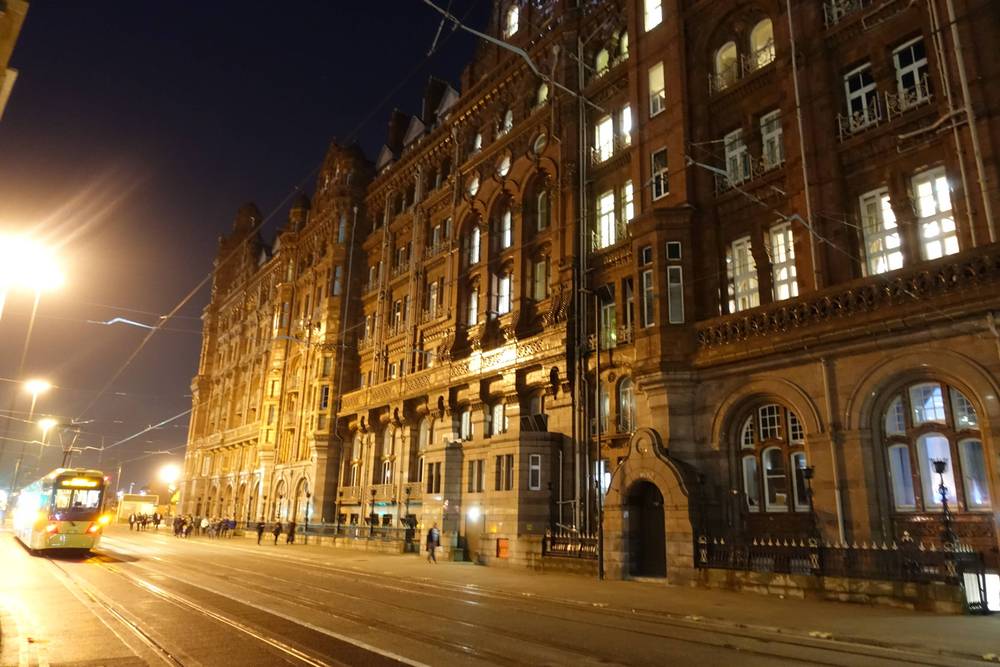 An ornate red-brick Victorian hotel building is lit up on a Manchester street at night, with a tram passing.