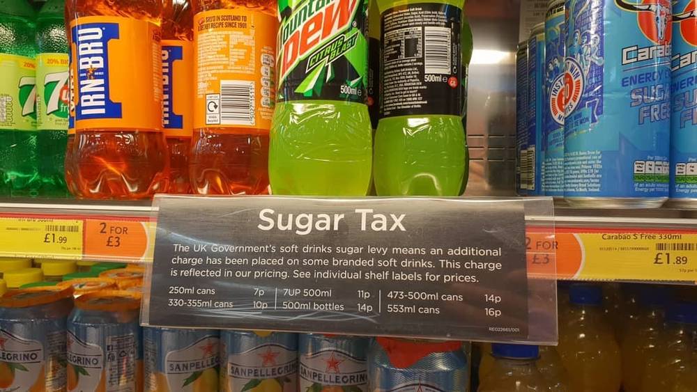 Supermarket drink shelf with a sign explaining the UK sugar tax on soft drinks.