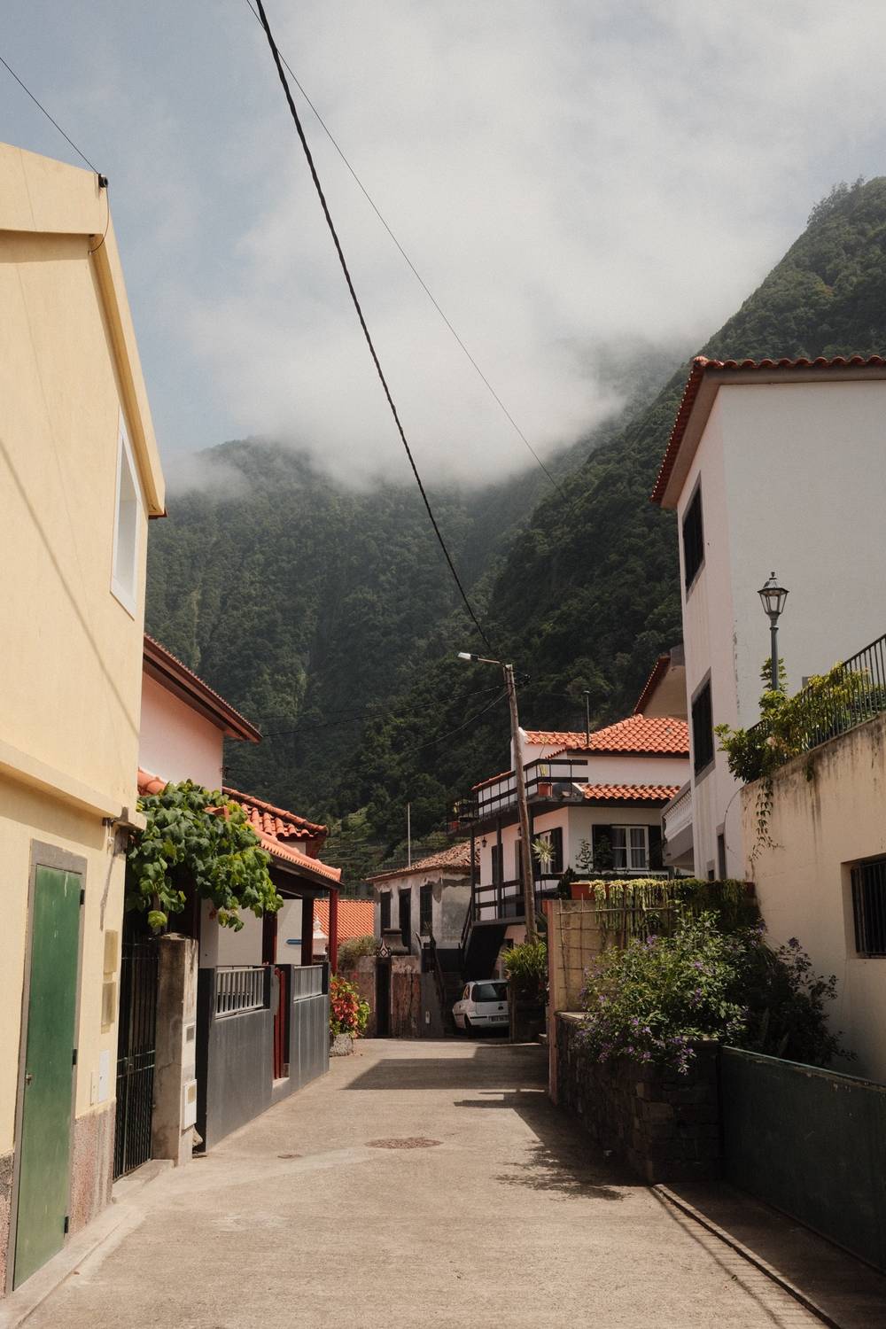 A quiet narrow street lined with whitewashed buildings leads toward a towering cloud-covered mountain in a village on Madeira, Portugal.