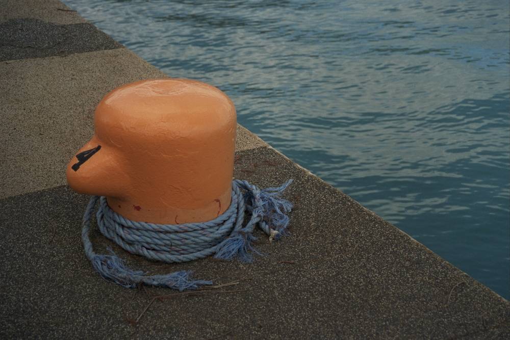 An orange-painted mooring bollard marked "4" on a concrete dock edge, wrapped at its base with frayed pale-blue rope, beside calm blue-green water.