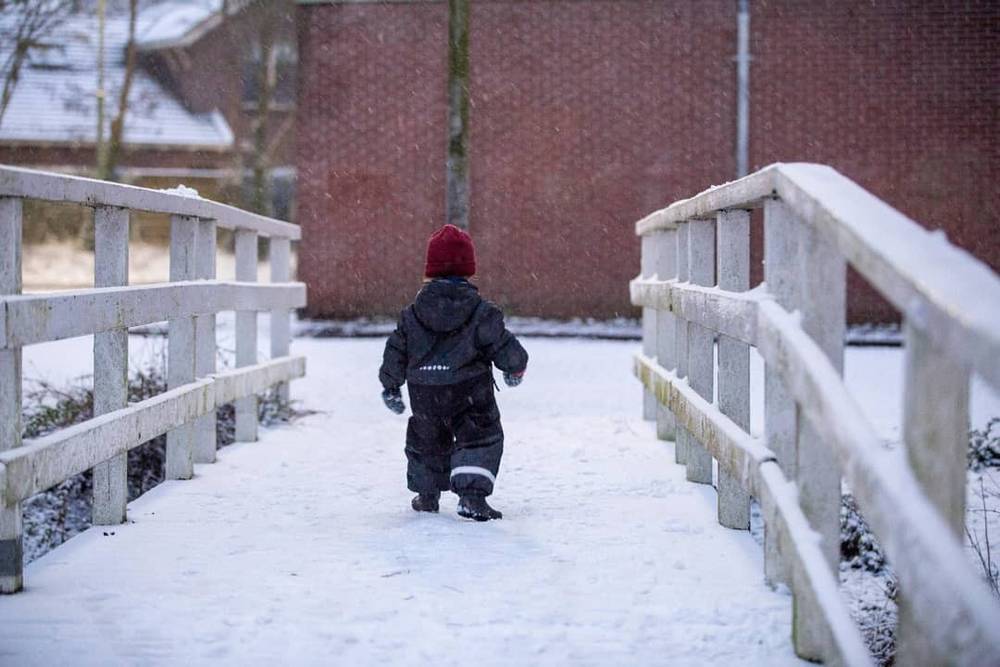 A child in a red hat walking across a snow-covered wooden bridge.