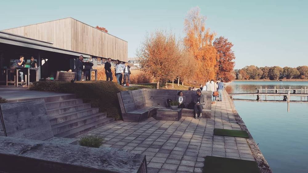 People gathered outdoors beside a lake at a conference venue with wooden buildings.