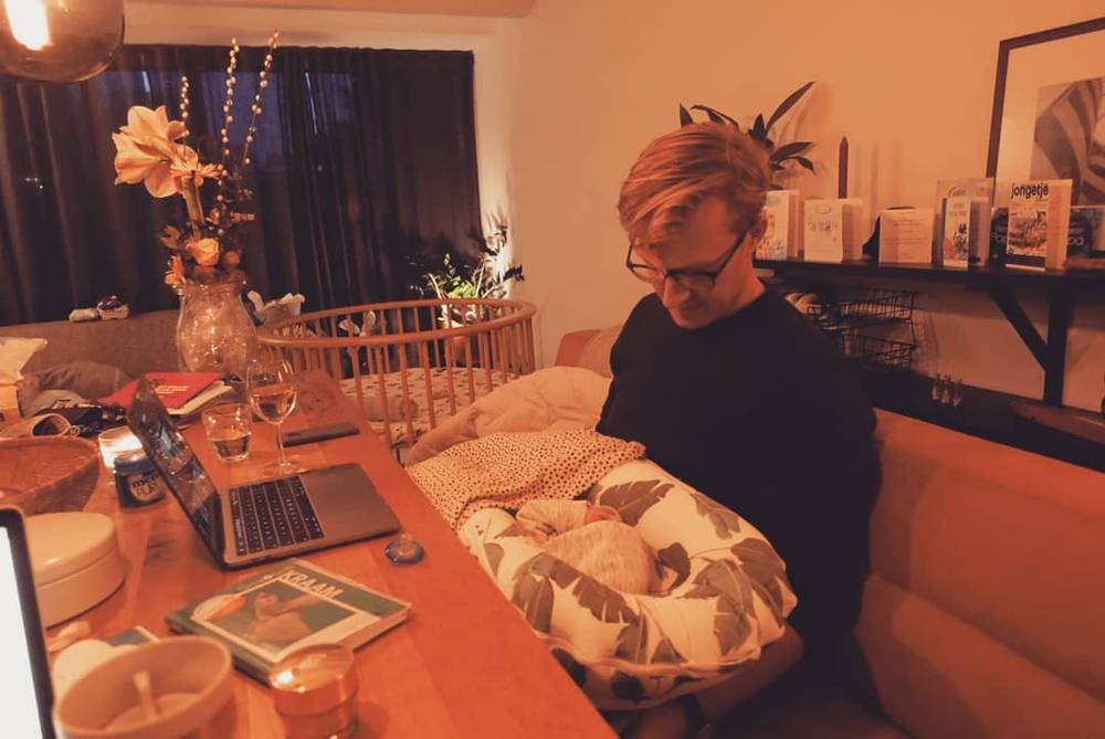 A man sits at a desk with a laptop beside a wicker moses basket holding a sleeping newborn, warm indoor lighting.