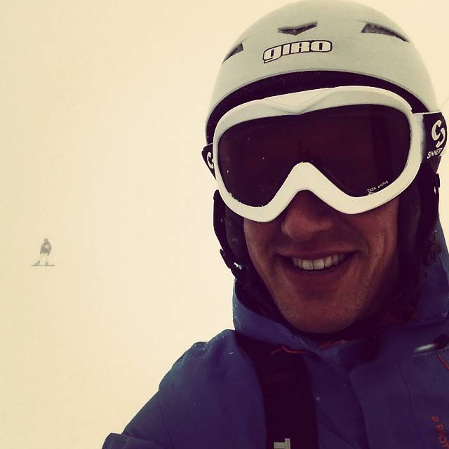 A smiling man wearing a white ski helmet and goggles, selfie style, with a snowboarder visible in the background.