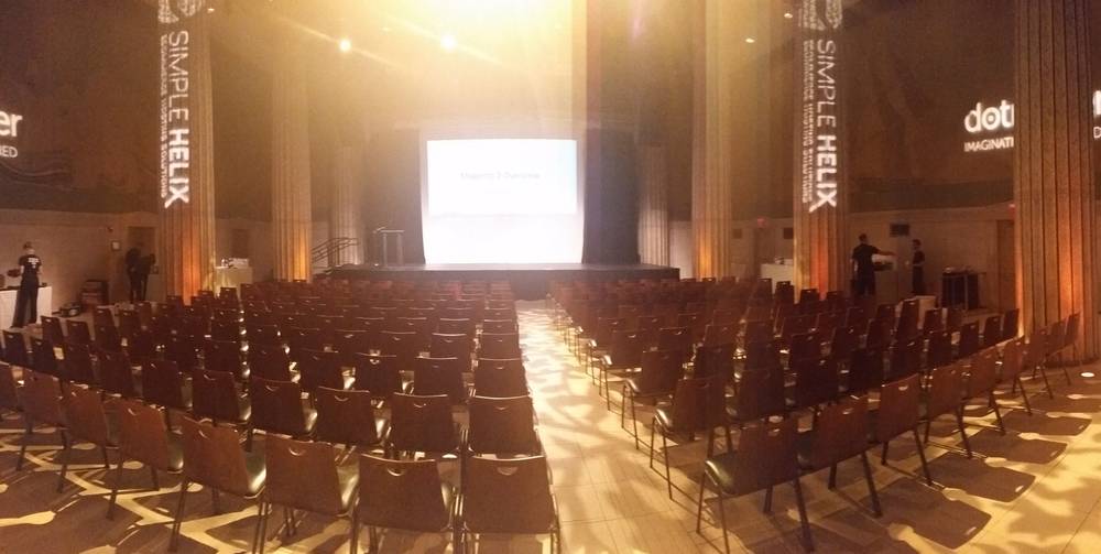 Rows of chairs fill a columned hall beneath a projection screen for a conference session.