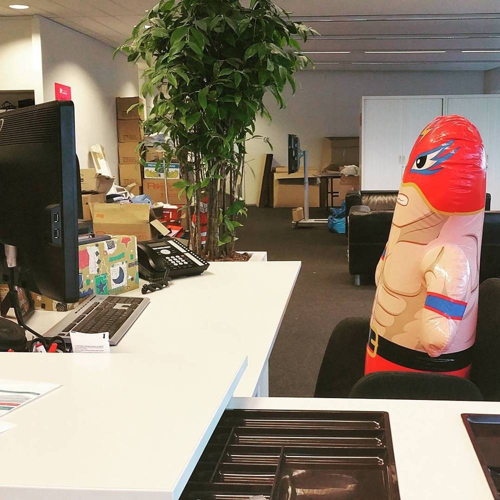 An inflatable wrestler figure standing on an office desk near computer monitors.