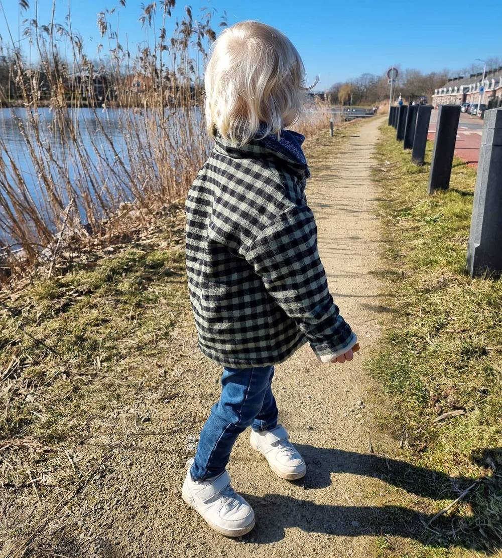 A child in a black-and-white checked jacket walking along a path beside a lake.