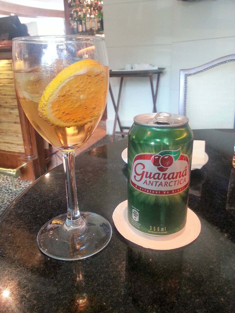 A glass of wine with an orange slice next to a can of Guarana Antarctica soda on a dark table.