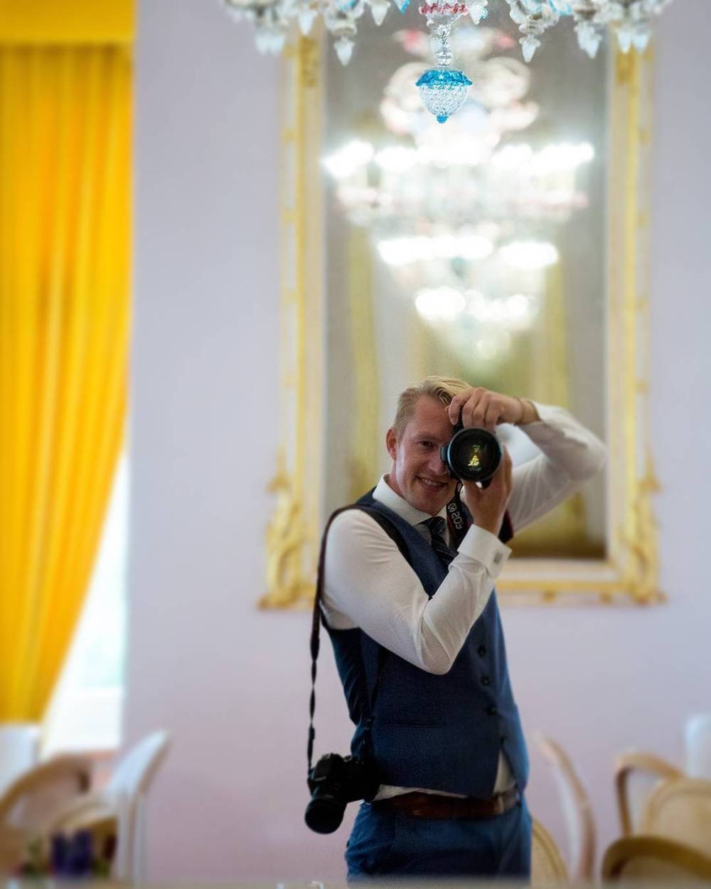 A man in a waistcoat photographs his reflection in an ornate gilded mirror.