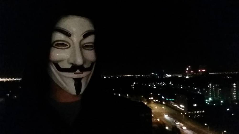 A person wears a Guy Fawkes mask for a nighttime selfie with city lights in the background.
