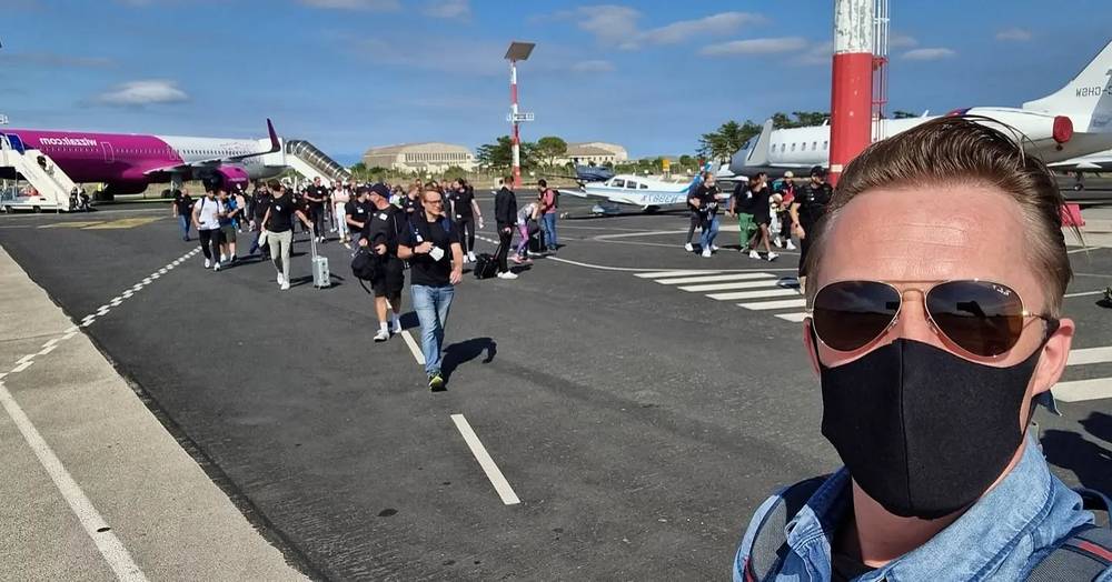 Guido wearing a face mask in the foreground, with a group of travelers walking across an airport tarmac toward small planes.