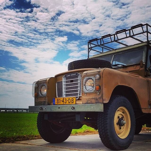 A vintage orange Land Rover with a roof rack parked on a road under a cloudy sky.