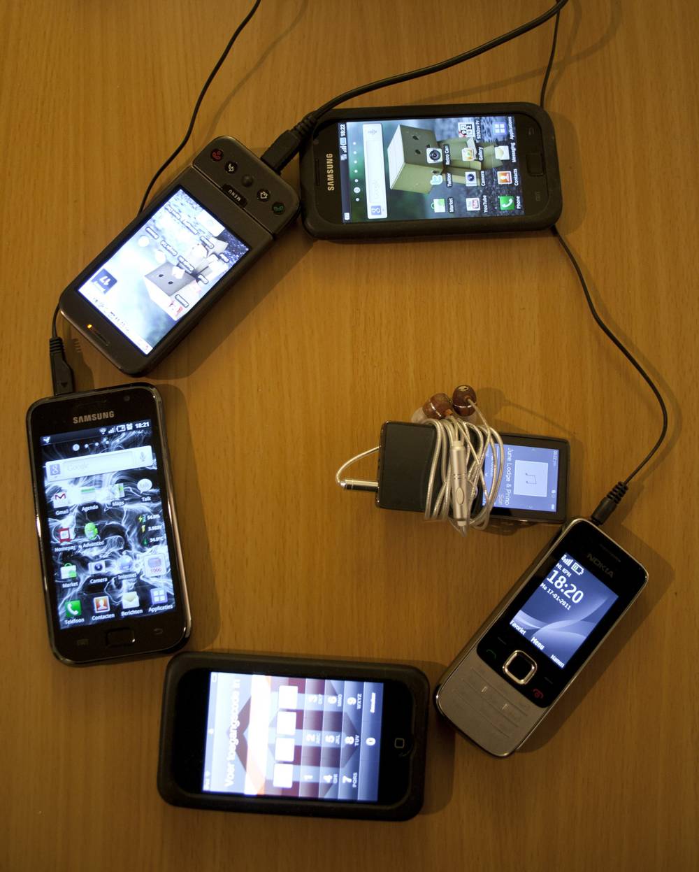 Five phones and mobile devices are arranged in a circle, all connected by charging cables.