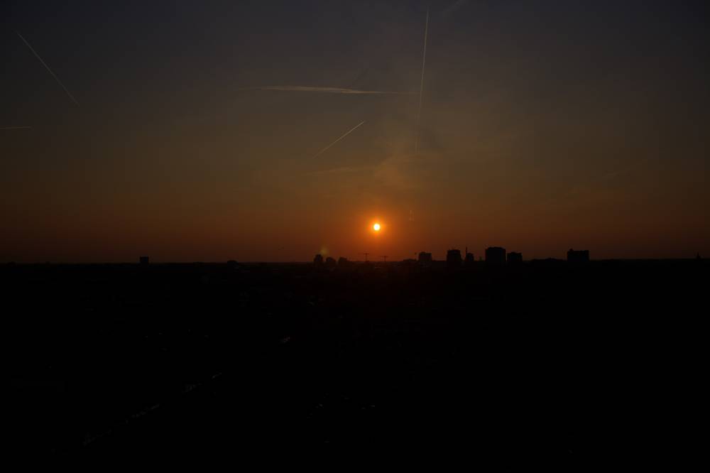 A small sun glows low over a dark, distant city skyline at dusk.