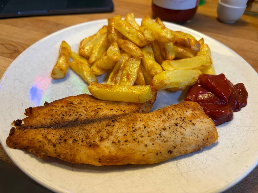 ein weisser teller mit einem braun geröstetem tilapia-filet und selbstgemachten, leicht glänzenden pommes aus dem airfryer. daneben ein grosser klecks ketchup.