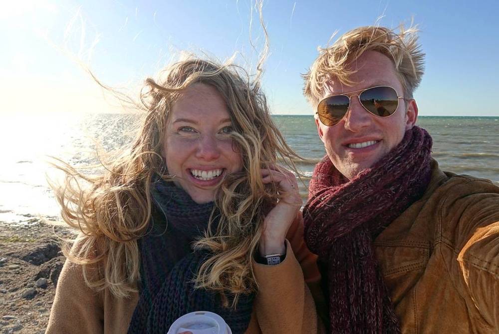 A couple smiles for a windswept selfie by the sea, hair blowing in the wind.