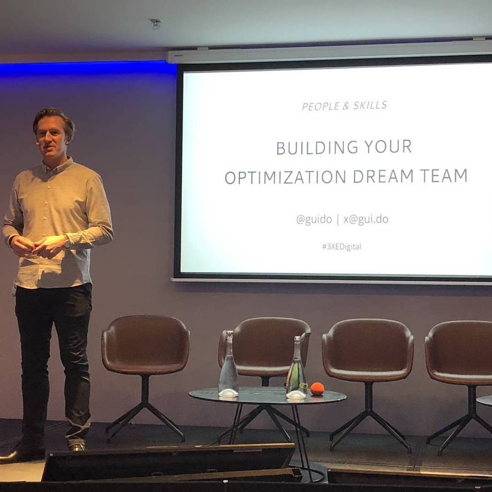 A man stands on stage beside a screen titled "Building Your Optimization Dream Team", empty chairs behind him.