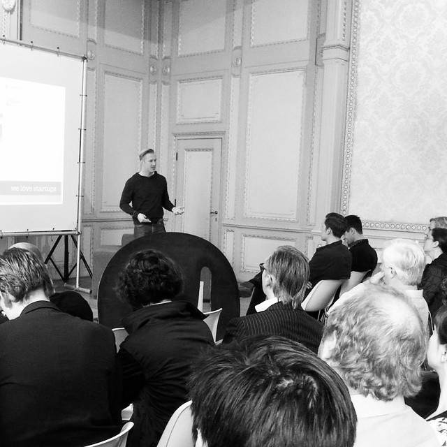 A man presents to a seated audience in a black-and-white photo, standing beside a screen.
