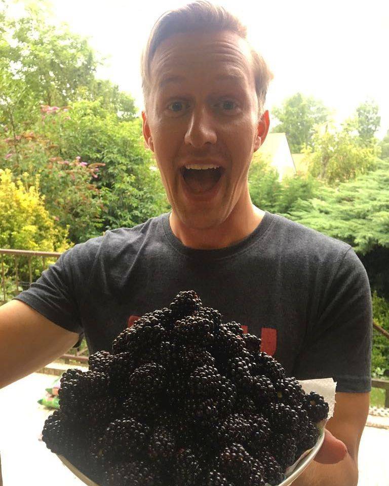A man holding a large mound of freshly picked blackberries, smiling in a garden.