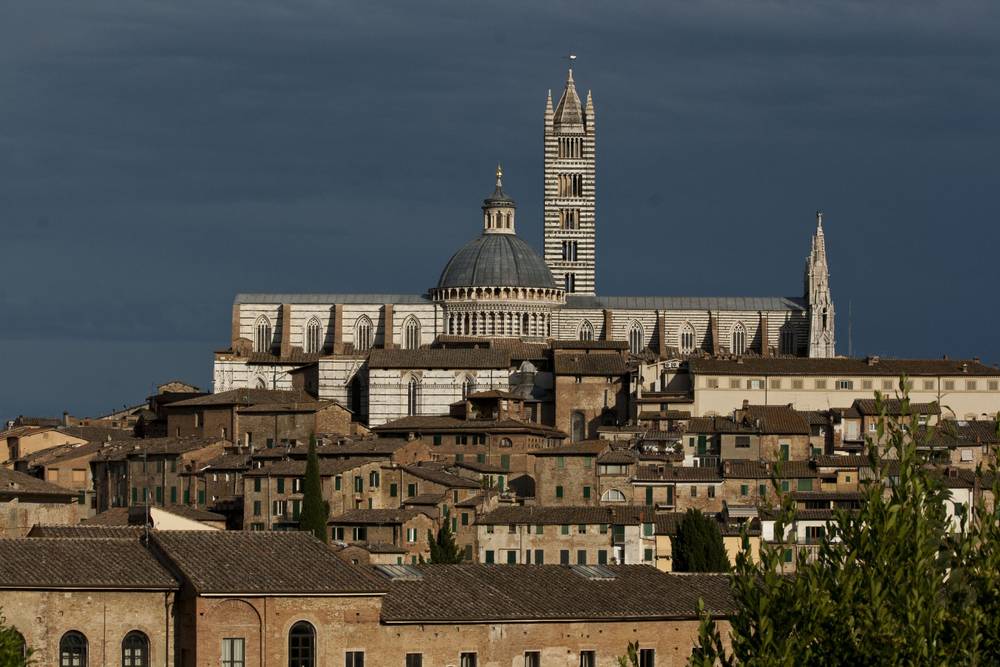 Siena's cathedral and surrounding rooftops glow under a stormy sky.