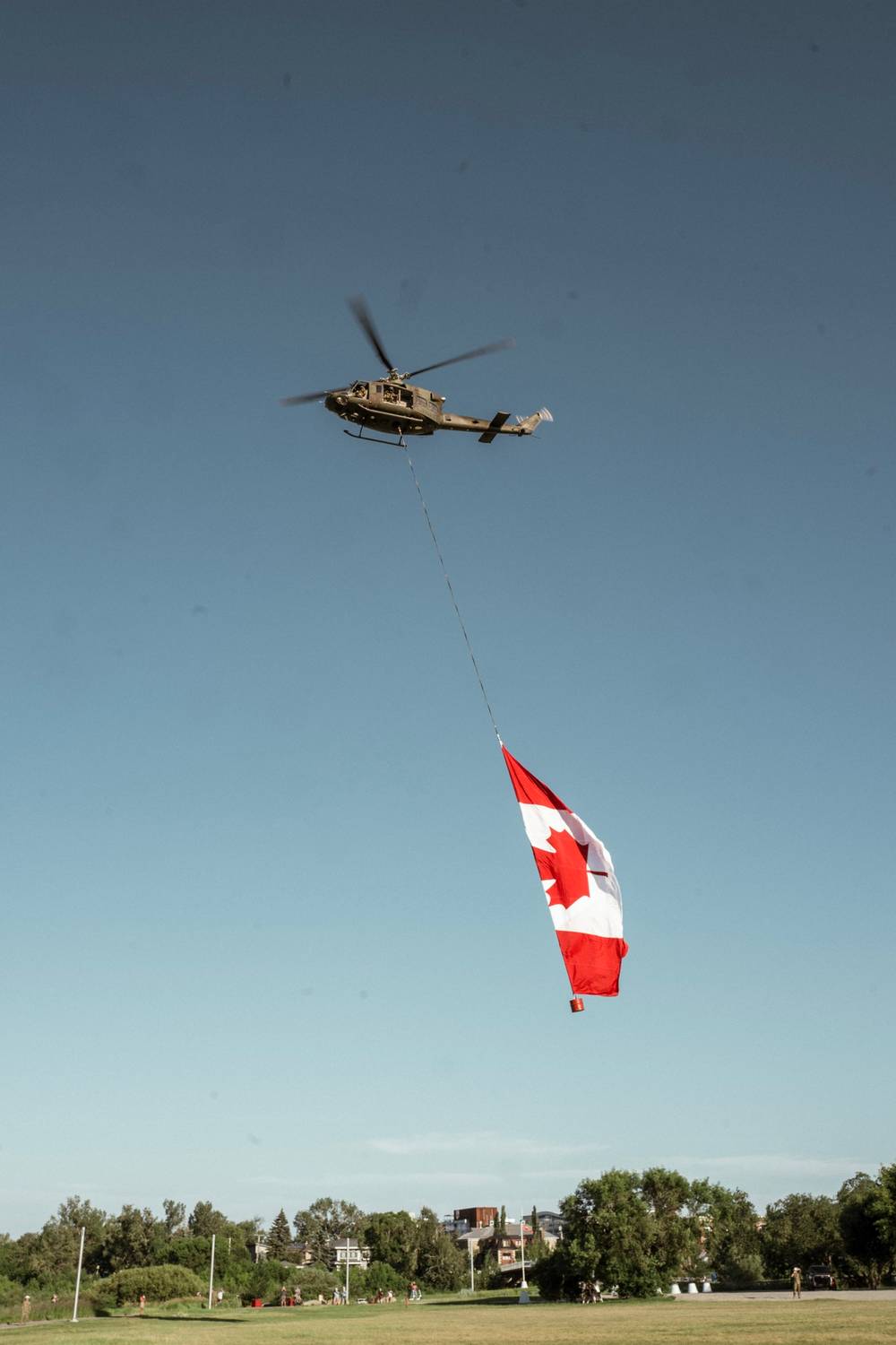 A photo of a military helicopter flying overhead against a blue sky with a large Canadian flag flying from a rope attached to the helicopter