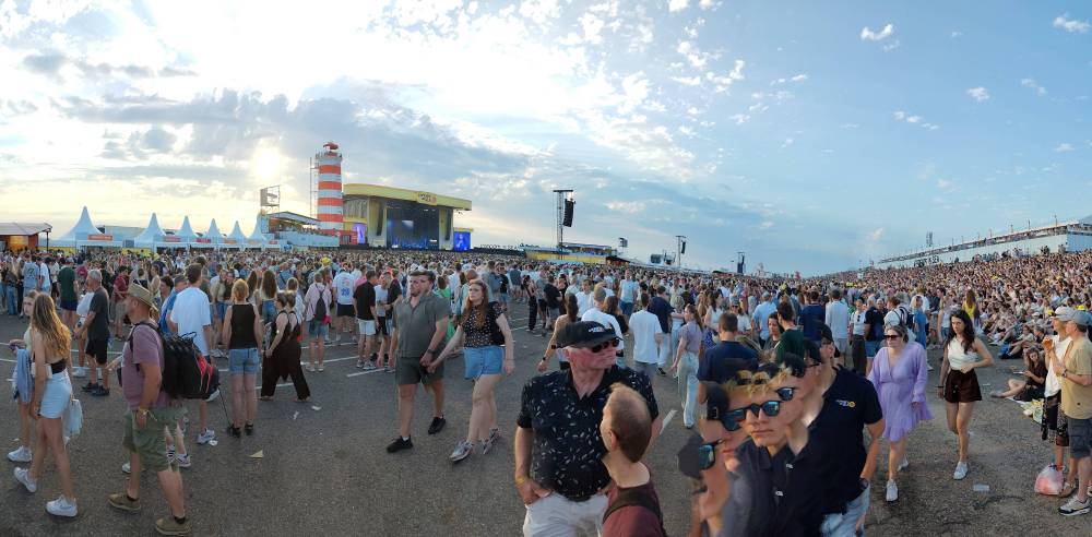 A festival crowd gathers near a lighthouse-shaped stage tower under a bright sky.