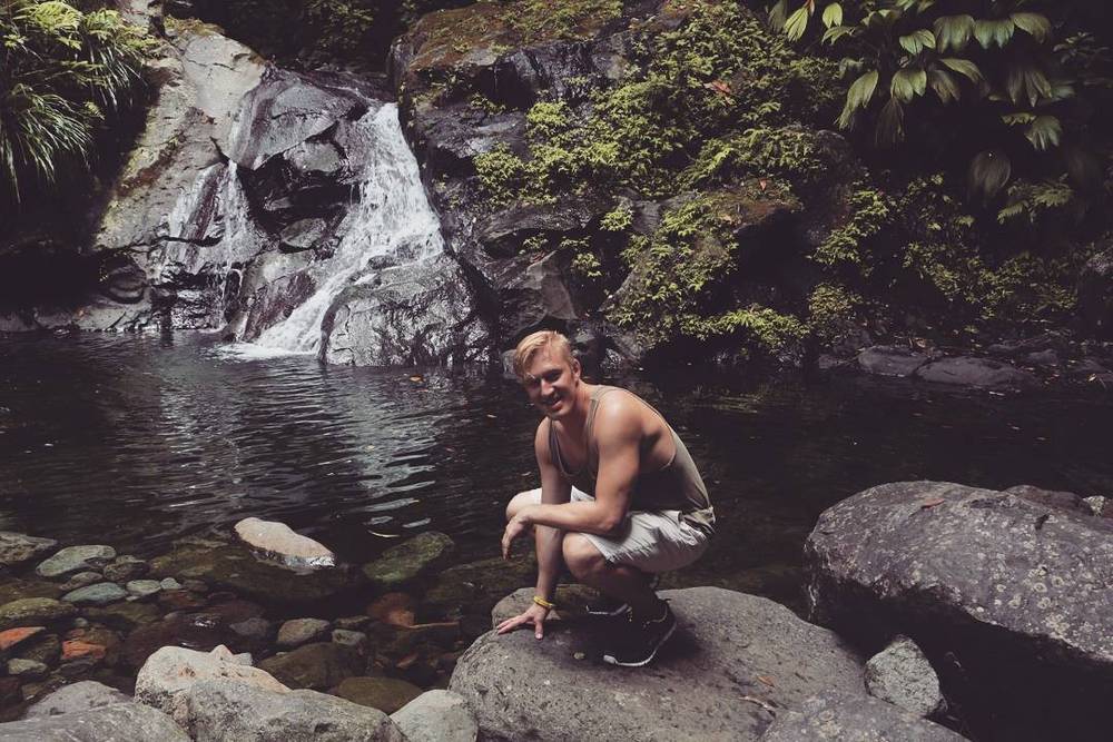 A man crouching on rocks beside a small waterfall pool in a forest.