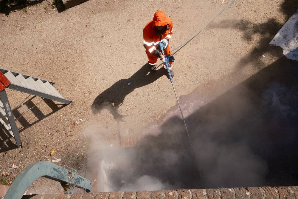 Man in Amsterdam maintenance clothes pressure washes a wall below the camera man with a ladder off to his left