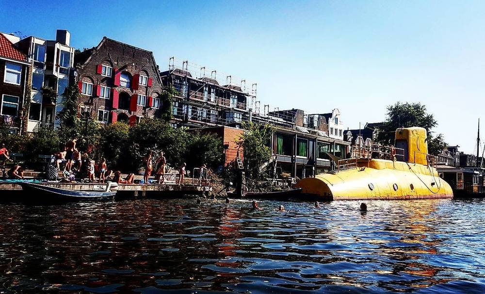 People swimming near a yellow submarine-shaped houseboat on an Amsterdam canal lined with old buildings.