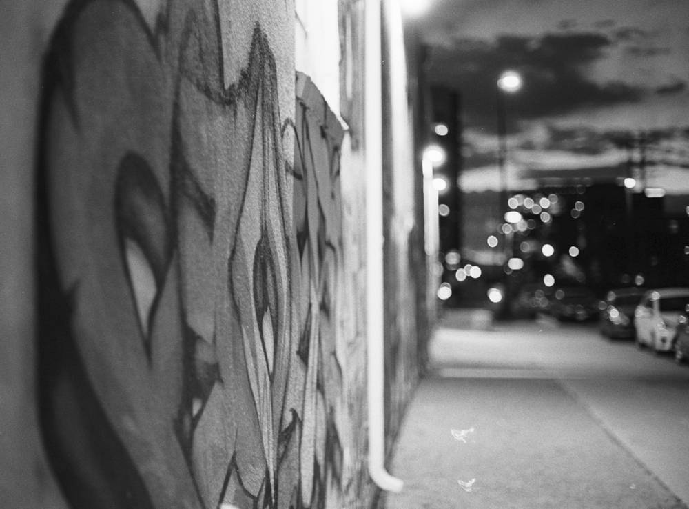 Night street scene with a graffiti covered wall in sharp focus in the foreground and out of focus streetlights, parked cars, and a moody sky in the background.