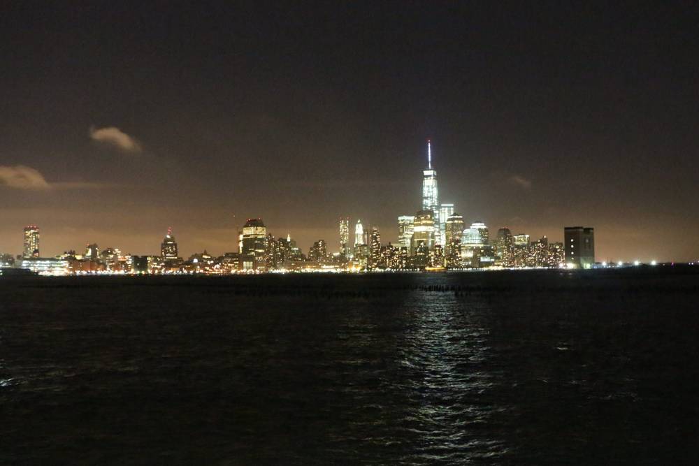 The Manhattan skyline, including One World Trade Center, glows across the water at night.