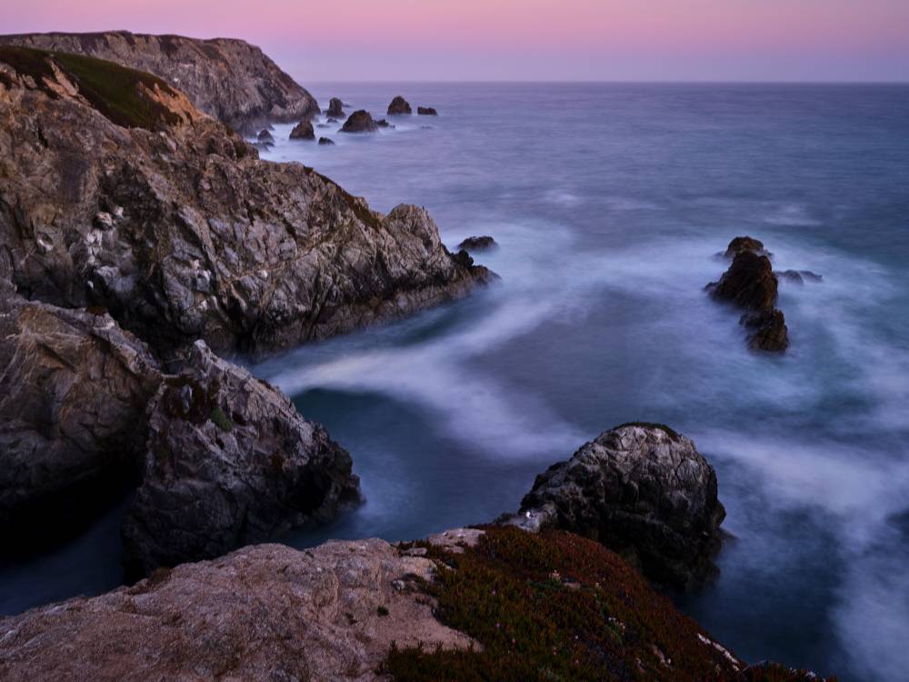 Long exposure of a rocky cove at dusk. Dark craggy cliffs frame silky white water swirling around sea stacks, with a pink and lavender sky fading to the horizon.