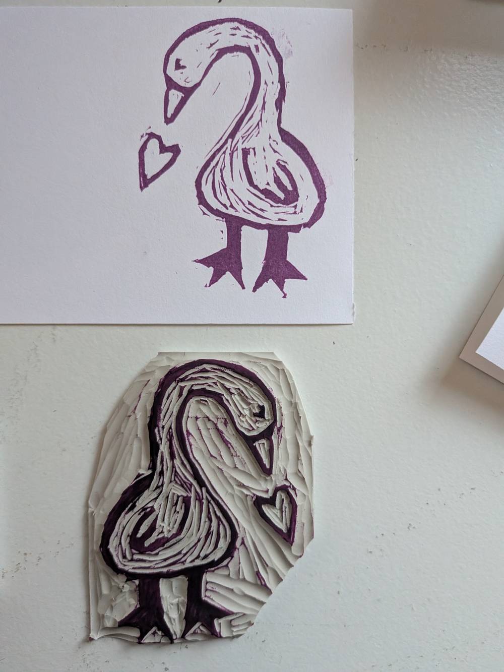 Below a linocut outlineof a mother goose in profile with textured body bending over with a heart below its beak. 
Above: a roughly cut card with print of mother goose. 

The two geese face in opposite directions 