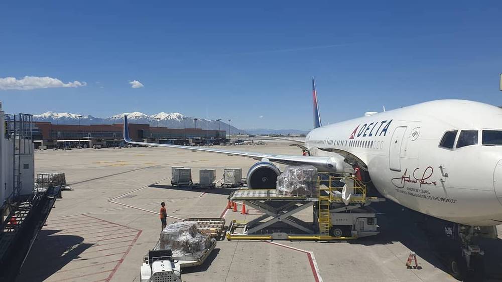 A Delta plane sits on the tarmac with mountains in the background as ground crew load luggage.