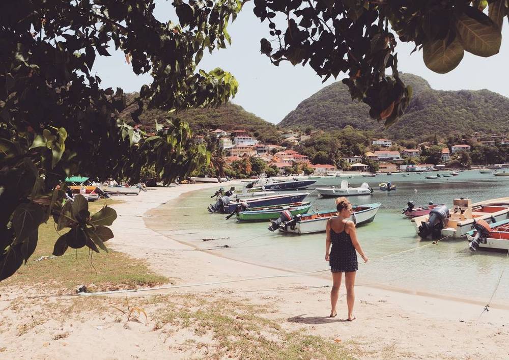 A woman standing on a beach overlooking a bay with boats and hills.