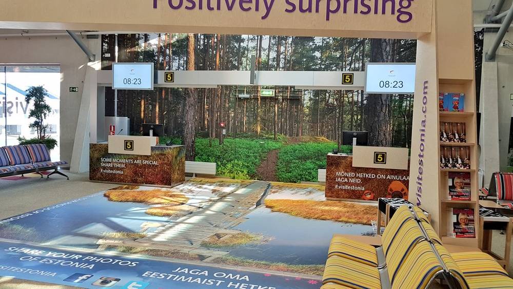 An airport terminal displays a "Positively surprising" tourism ad for Estonia with a forest scene.