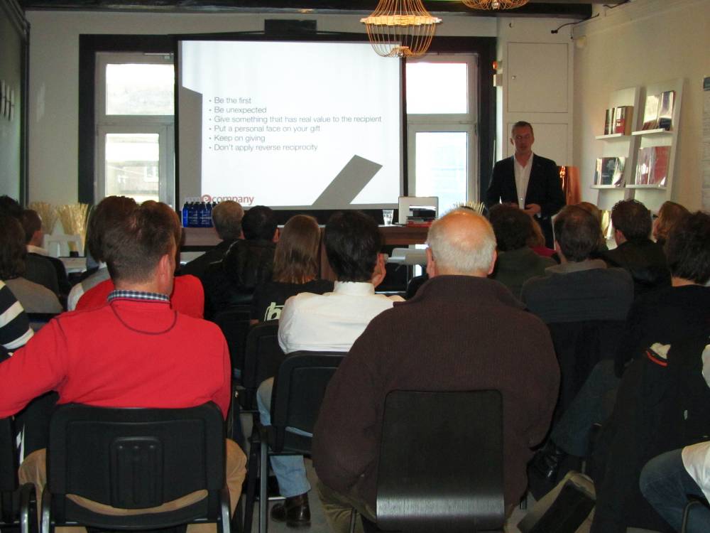 A speaker addresses a seated audience in front of a slide about giving unexpected gifts.