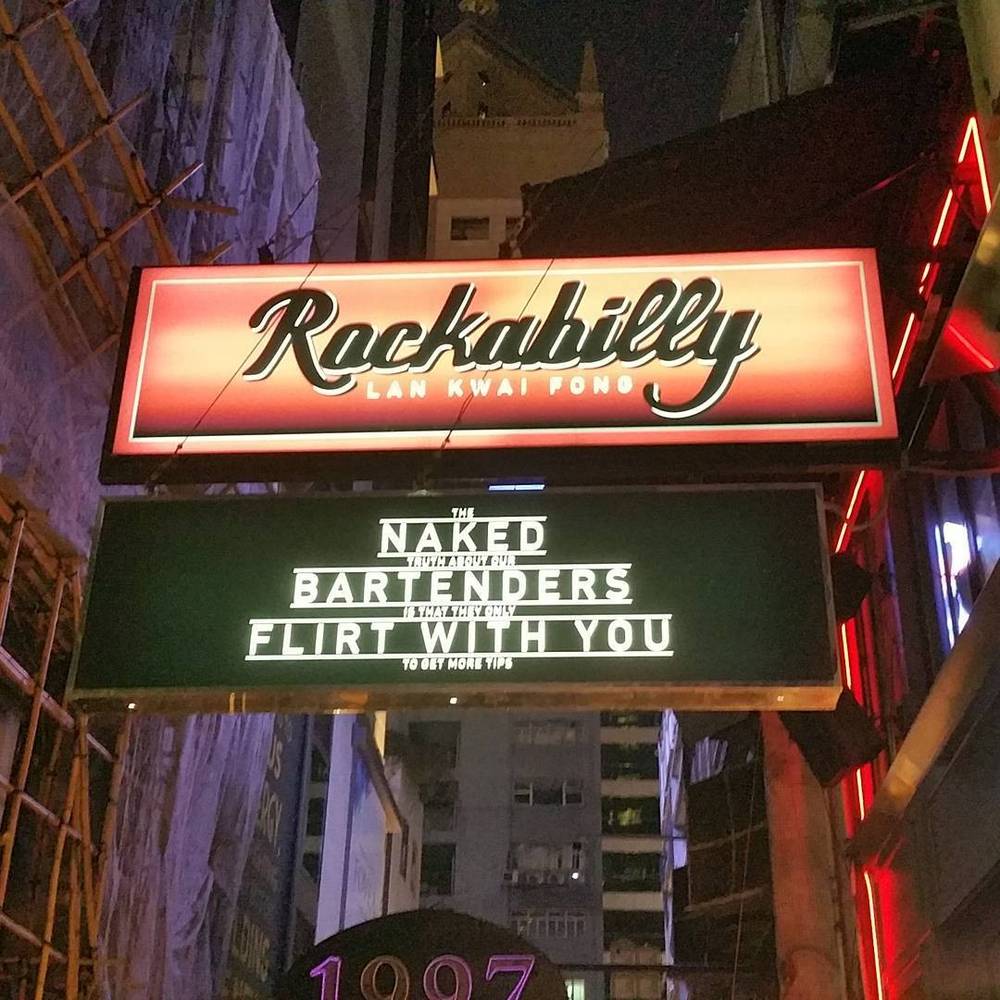 A neon "Rockabilly" bar sign in Lan Kwai Fong, Hong Kong, above smaller signs reading "The Naked Bartenders" and "Flirt With You".