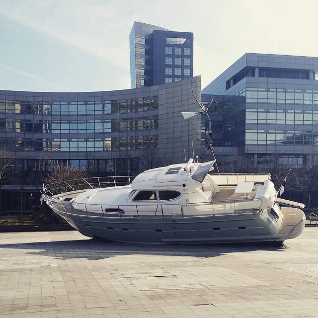 A boat displayed on a paved plaza in front of a modern glass building, with no water nearby.
