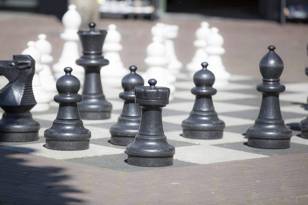 Large black and white chess pieces stand on an outdoor pavement chessboard.