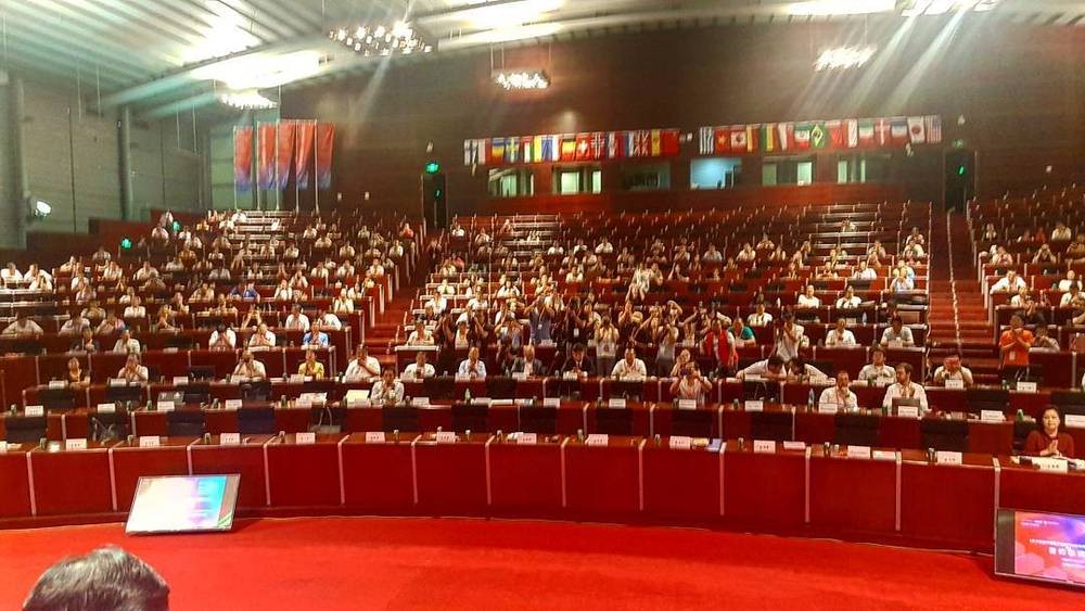 A large tiered auditorium filled with attendees, flags of many countries lining the walls.