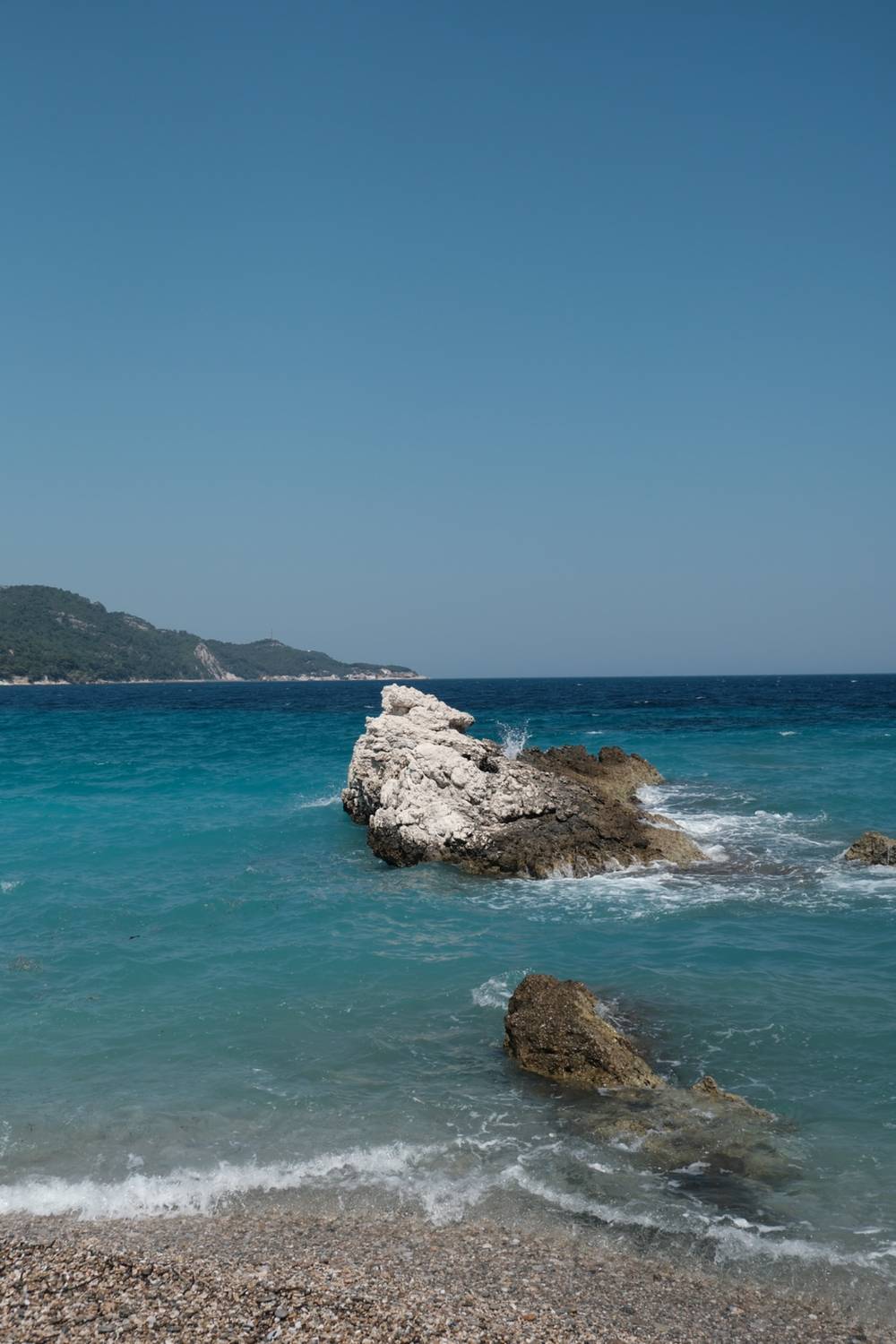 a jagged rock formation surrounded by splashing turquoise waves, Kokkári's beach in the foreground and green hills across the water.