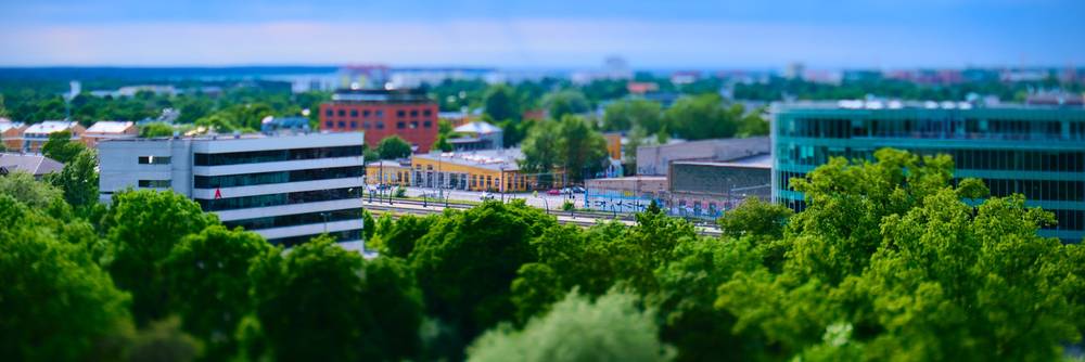 A tilt-shift panoramic photograph of an urban landscape, creating a miniature model effect. Lush green trees fill much of the frame, with prominent modern buildings in white and teal visible through the dense foliage under a clear blue sky.