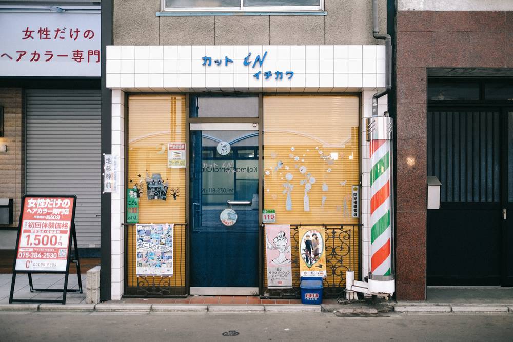 Another barber shop with white walls made of square tiles, their name in blue (I rather like the typeface), warm yellow blinds down in the windows