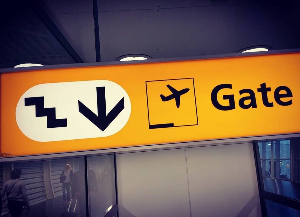 An orange airport signage panel pointing toward stairs, an escalator, and the departure gates.