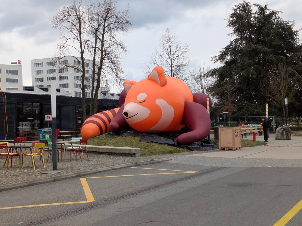 A giant inflatable red panda, the mascot of Japan Impact, appears to be sleeping peacefully on a small grassy hill partially covered with a black tarp. The large orange and maroon figure has a striped tail and closed eyes. It is situated in an outdoor plaza next to a patio area with colorful cafe tables and chairs. In the foreground is a paved road with yellow line markings, and in the background are bare winter trees, modern white buildings, and an overcast sky.