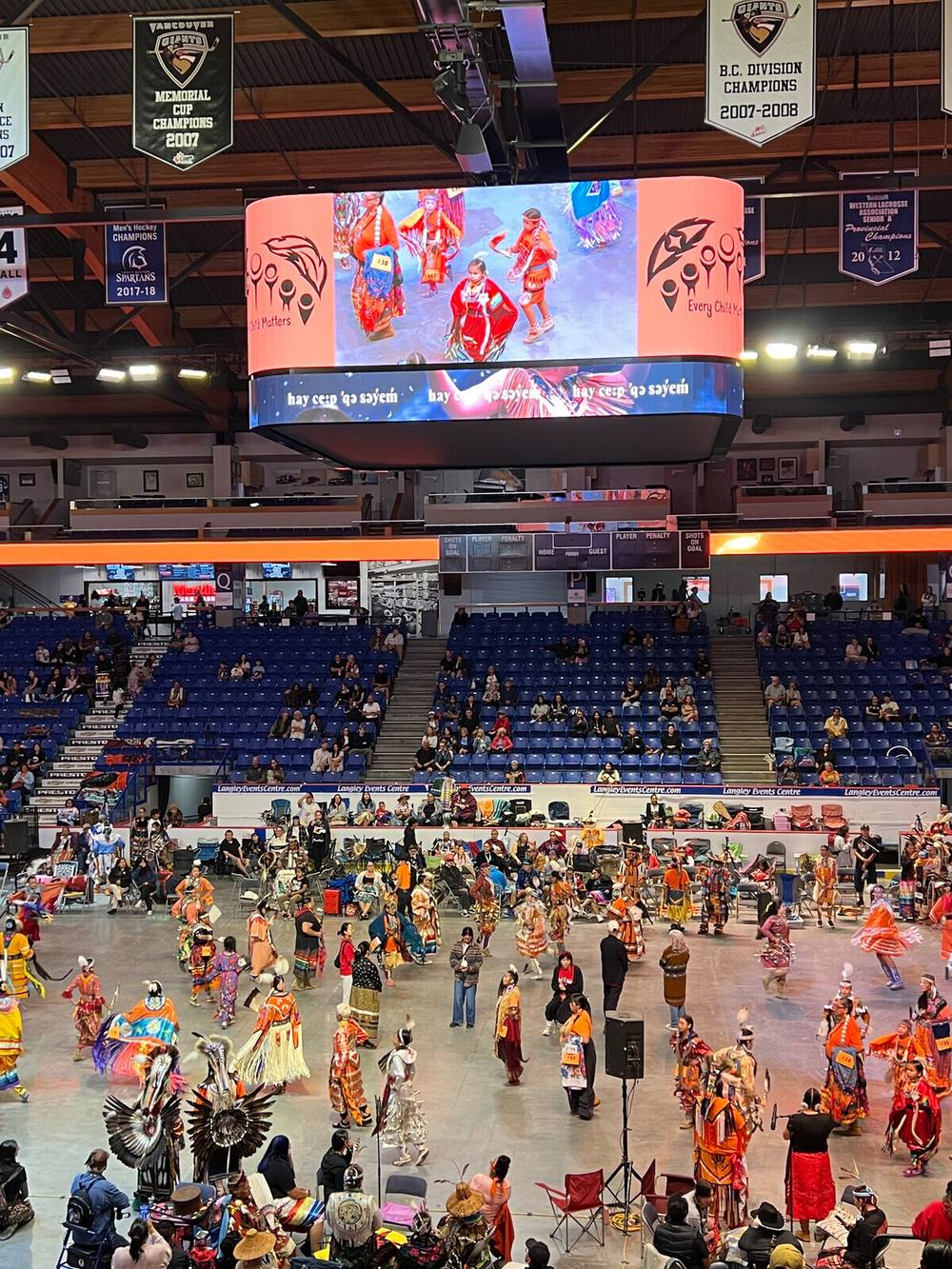 A large group of people dressed in colorful traditional Indigenous attire are participating in a powwow inside an arena with spectators seated in the stands.