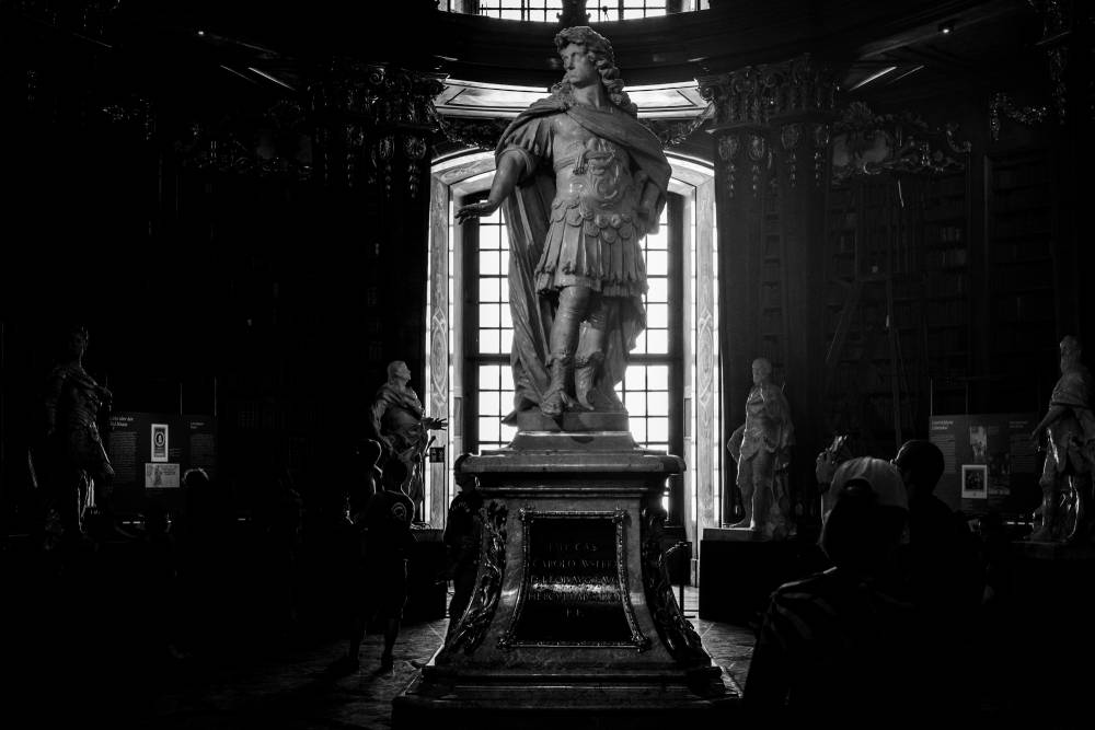 A dramatic, low-angle black and white photograph of a large marble statue depicting a powerful figure, possibly a Roman emperor. The figure is silhouetted against a tall arched window in a grand, ornate hall, which appears to be part of the Vienna State Hall, with bookshelves visible in the deep shadows. Dark human figures are seen in the foreground, looking up. The entire image has a textured, printed look.