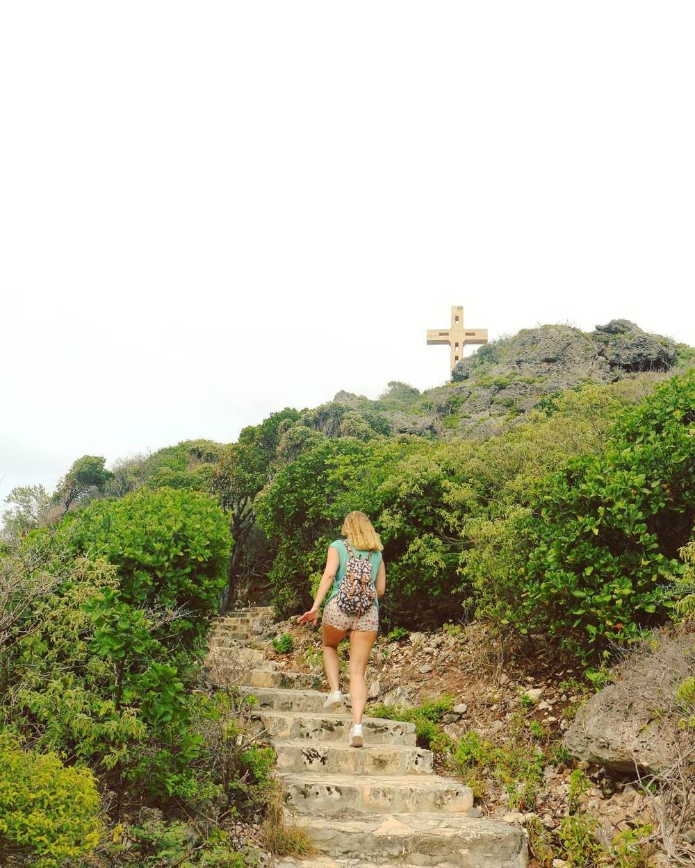 A woman walking up stone steps toward a cross on a hilltop, surrounded by greenery.