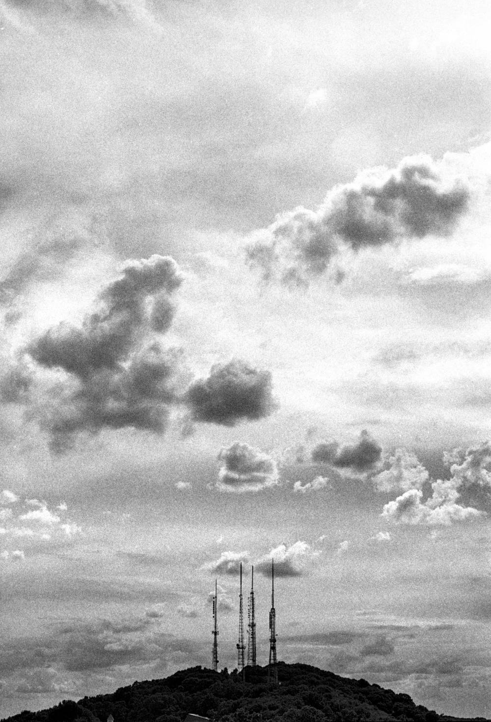 Distant view of Pinnacle Hill in Rochester, NY, seen from Cobbs Hill. A cluster of tall broadcast antenna masts rises from the wooded crest of the low hill along the bottom of the frame. Above, a dramatic sky fills most of the image: broken cumulus clouds pile up in bright and dark banks against patches of clear sky.